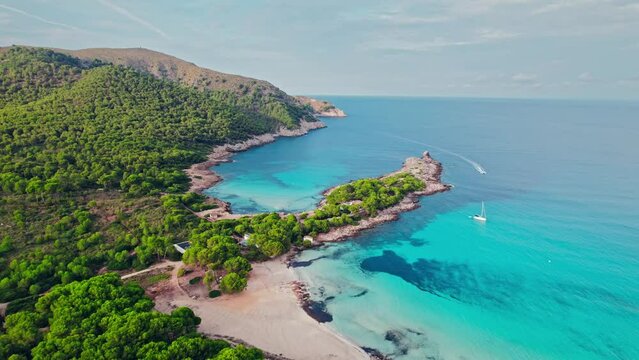 Vessel sailing by a Beautiful tropical green rocky coast with turquoise waters. Paradise alike bay in Cala Agulla, Mallorca, Balearic Islands, Spain with a tourist boat exploring the location.
