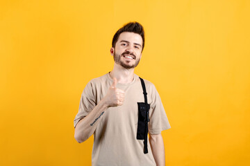 Cheerful young man wearing t-shirt posing isolated over yellow background doing happy thumbs up gesture with hand. Looking at the camera showing success.