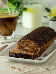 Chocolate cake roll dessert with cut piece and coffee with milk on white plate