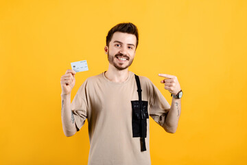 Cheerful young man wearing t-shirt posing isolated over yellow background pointing finger at credit or debit card. Shopping and finance concept.