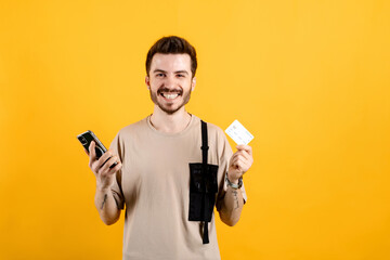 Happy casual man wearing t-shirt posing isolated over yellow background holding credit card and mobile phone while smiling and looking at the camera.