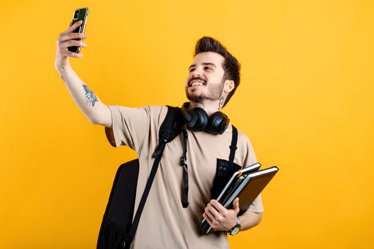 Young man smiling confident wearing beige tee posing isolated over yellow background university college concept. Doing selfie shot on mobile phone.