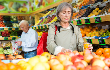 Old lady choosing mandarins while standing in salesroom of greengrocer. Old man shopping in...