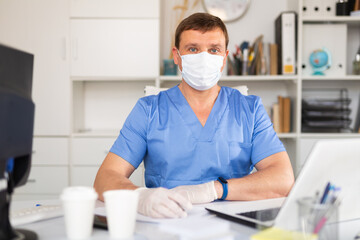 Fototapeta na wymiar Confident male doctor in blue medical uniform, mask and gloves working on laptop in clinic office