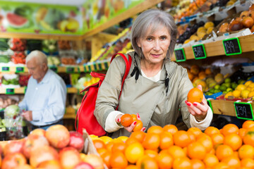 Fototapeta premium Mature woman selecting tangerines in supermarket. Senior man shopping in background.