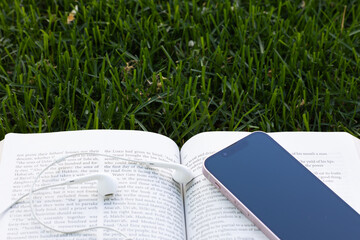 Open bible with mobile phone and headphones laying on the grass with copy space