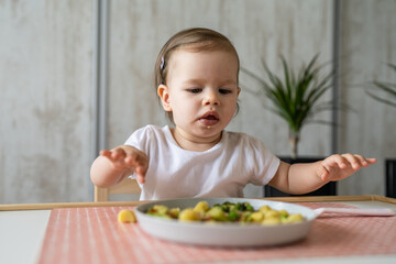 One baby girl small caucasian toddler eating while sit at the table alone at home copy space little child having lunch healthy food meal in room in day front view
