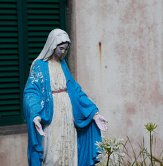 old weathered garden statue of virgin mary reaching hands out in blue and white in park with flowers religious garden gnome  outdoor garden decoration in with flowers in foreground vertical format