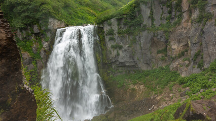 Fototapeta na wymiar The waterfall flows down from a sheer cliff. Green vegetation on rocky slopes. Kamchatka.