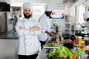 Head chef wearing cooking uniform while standing in restaurant professional kitchen with arms crossed. Gastronomy expert cutting fresh vegetables and mixing spices for dinner service.