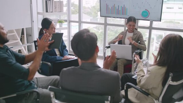 Confident business woman in wheelchair presenting during business meeting with colleagues. Asian business team celebrating success together in boardroom.