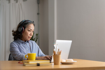 Mature asian woman wearing headphones using laptop, making video call, sitting at table in kitchen,...