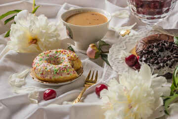 Breakfast in bed with various delicious glazed donuts on the plate, cup of coffee and sweet cherry berries. Side view.