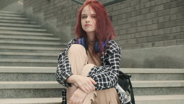 Portrait of joyful carefree girl student with long red hair sitting in city on stone staircase. Happy teen hipster female with headphones listening to favorite music online audio audio enjoying song