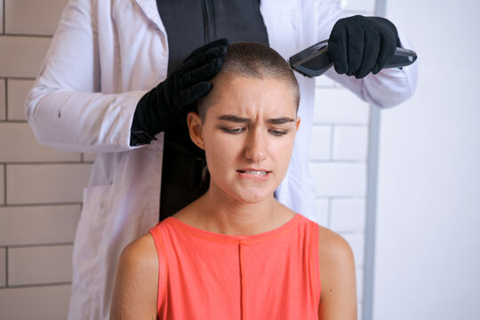Young woman is forcibly shaving her head with an electric razor, holding typewriter in black gloves, a girl with a sad look with tears in her eyes. Close-up, copy space