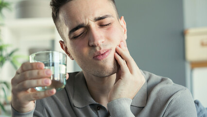 Young man with sensitive teeth and hand holding glass of water	