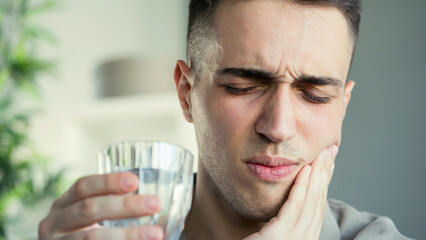 Young man with sensitive teeth and hand holding glass of water	
