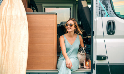 Happy young woman sitting with her boston terrier dog at the door of her camper van during a trip