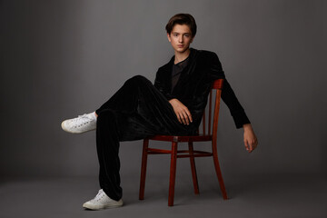 The guy poses in the studio in a black suit and white sneakers sitting on a chair on a gray background