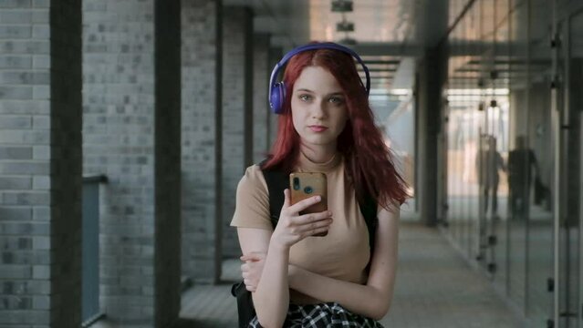 Young attractive girl in casual clothes is standing outdoors in city with headphones listening to music on modern phone