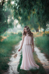 A blonde with beautiful curls in a long evening dress walks in the park in the evening. A beautiful girl with curls of hair and evening make-up holds twigs of reeds in her hand.