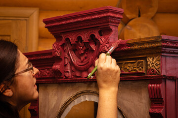 Attentive lady coloring massive wooden cupboard with astounding carved ornaments in red with brush...