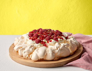 food, culinary, baking and cooking concept - close up of pavlova meringue cake decorated with red berries on wooden serving board and kitchen towel over yellow wall background