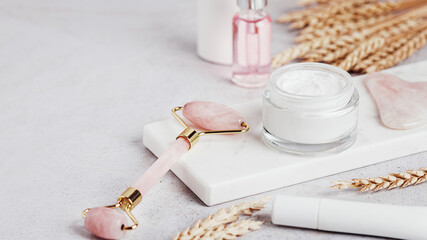 Facial roller, gua sha, serum and moisturizing cream on marble background