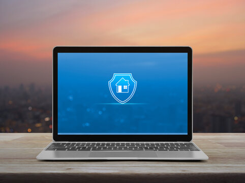 Home with shield flat icon on modern laptop computer monitor screen on wooden table over blur of cityscape on warm light sundown, Business home insurance and security online concept
