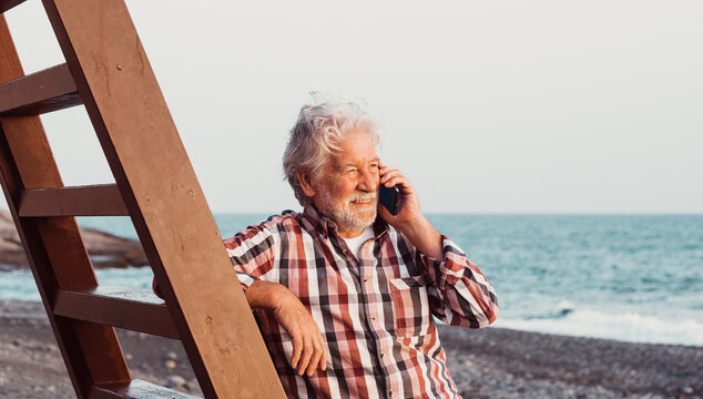 Smiling caucasian senior man on the beach looking at the golden hour of sunset at sea while talking on mobile phone, bearded male enjoying free time, vacation or retirement - Powered by Adobe