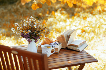 Bouquet of flowers, croissant, cup of tea or coffee, books on table in autumn garden. Rest in...