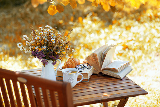 Bouquet of flowers, croissant, cup of tea or coffee, books on table in autumn garden. Rest in garden, reading books, breakfast, vacations in nature concept. Autumn time in garden on backyard