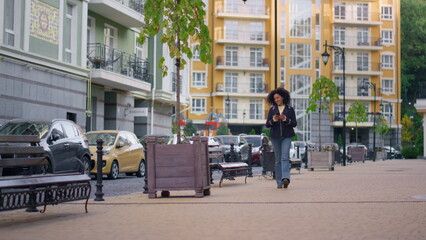 Joyful woman walking texting friends online. Modern residential area district.