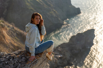 Woman tourist enjoying the sunset over the sea mountain landscape. Sits outdoors on a rock above the sea. She is wearing jeans and a blue hoodie. Healthy lifestyle, harmony and meditation