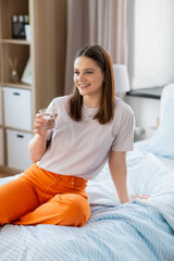 people concept - happy smiling girl with glass of water sitting on bed at home