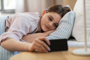 people, bedtime and rest concept - happy smiling teenage girl with smartphone lying in bed at home