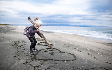 A girl in warm clothes and a hat with a backpack walks along the beach and draws drawings and symbols on the sand
