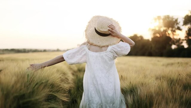 Back view of young girl happily walking in slow motion through a field touching with hand wheat ears.