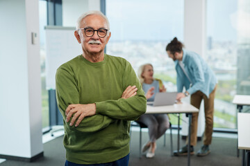 Fototapeta premium A proud senior student is standing in a classroom and smiling at the camera while educator teaching a woman in blurry background.