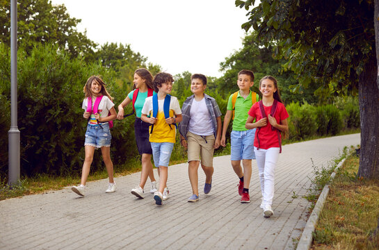 School friendship. Funny schoolchildren group with backpacks have fun walking together on path in park. Cheerful boys and girls in summer casual clothes return home from school lessons together.