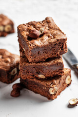 Stack of delicious brownie pieces with hazelnuts on marble background with knife, close up