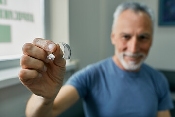 Senior man holding BTE hearing aid in hand on foreground, close-up. Treatment of deafness in...