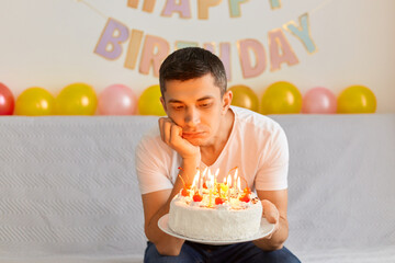 Indoor shot of young adult bored man feeling boring, having sad upset expression, sitting on sofa, waiting guests, celebrating birthday alone at home, wearing white t shirt.