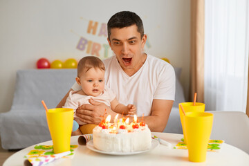 Portrait of brunette man with infant baby celebrating first birthday, sitting at table with birthday cake, father with excited facial expression hugging his infant daughter, celebrating together.