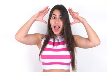 Obraz na płótnie Canvas young beautiful brunette woman wearing striped top over white wall looks with excitement at camera, keeps hands raised over head, notices something unexpected, reacts on sudden news.