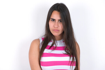 young beautiful brunette woman wearing striped top over white wall frowning his eyebrows being displeased with something.