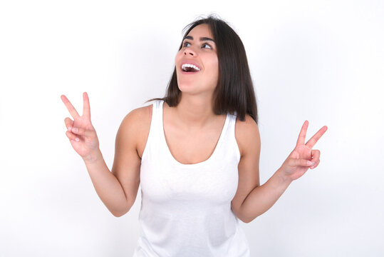 Isolated shot of cheerful Young beautiful brunette woman wearing white top over white wall makes peace or victory sign with both hands, feels cool.