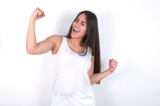 Attractive Young beautiful brunette woman wearing white top over white wall celebrating a victory punching the air with his fists and a beaming toothy smile