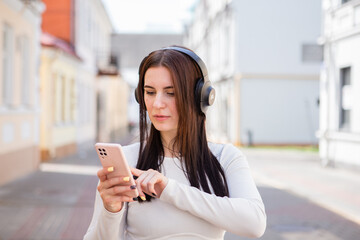 beautiful caucasian young cheerful woman in fashionable clothes listens to music using headphones and a smartphone and smiles while walking around the city