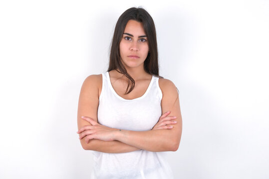 Picture of angry Young beautiful brunette woman wearing white top over white wall looking camera.